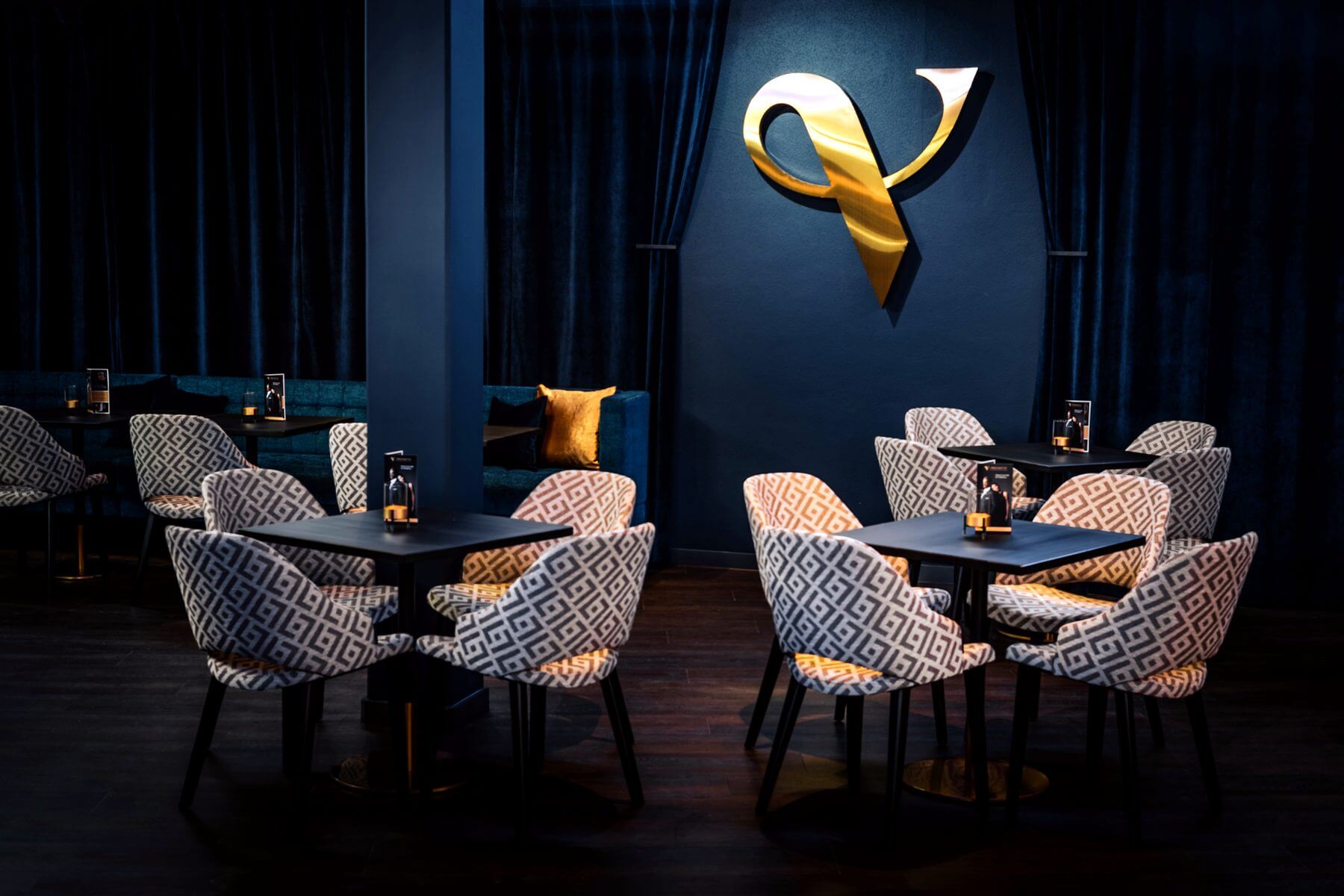 Lounge-Sitzgruppe im Zaubertheater VERSCHMITTST mit gemusterten Stühlen, kleinen Tischen und Logo im Hintergrund